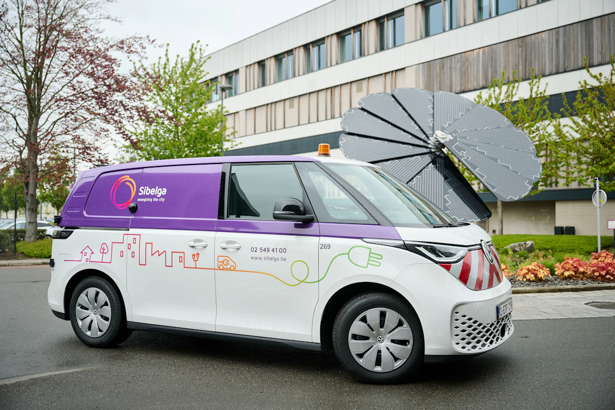 A purple and white Volkswagen ID. Buzz electric van parked in front of a commercial building with solar panels on the roof. The building has the logo for Sibelga, a Belgian energy company.